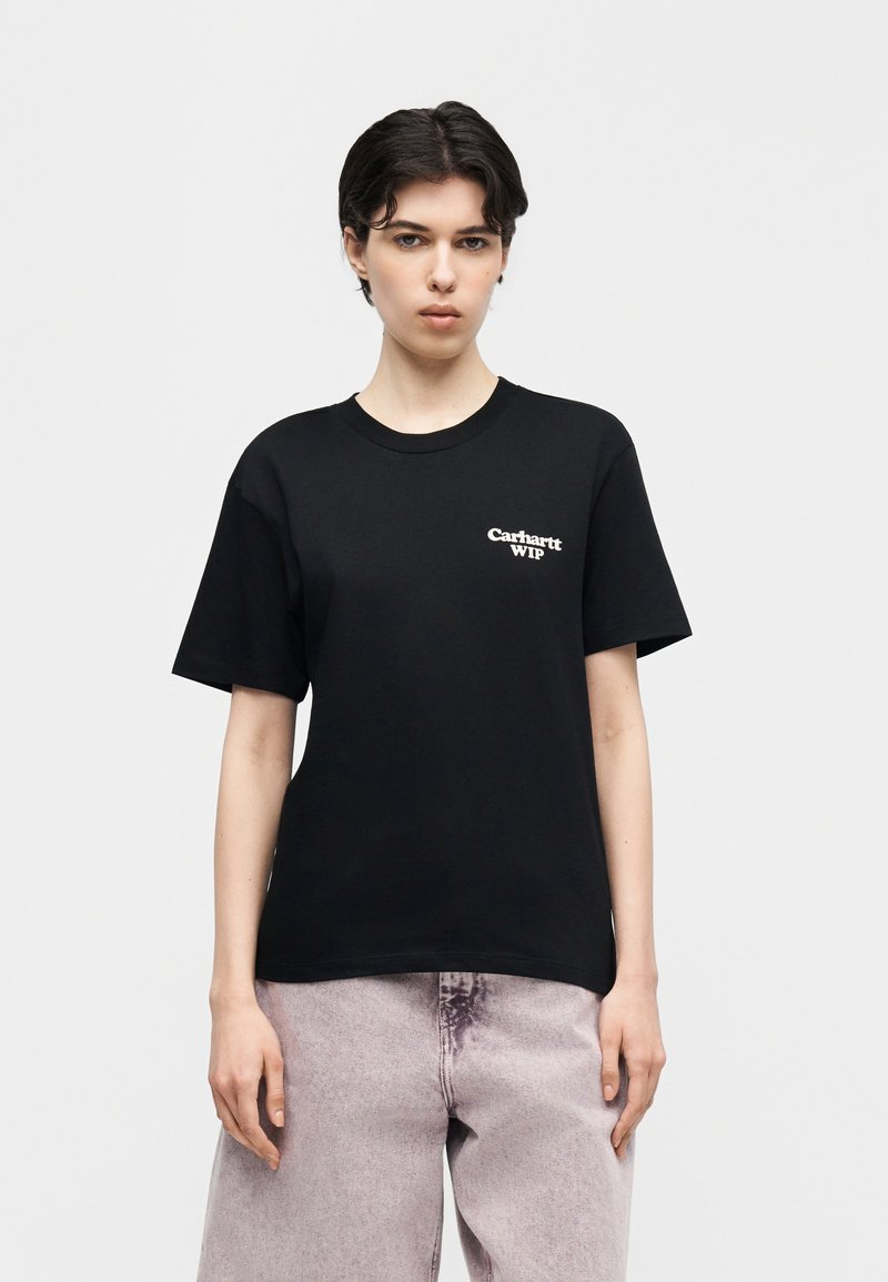 Person mit kurzen dunklen Haaren trägt ein schwarzes Carhartt WIP T-Shirt und hellviolette, gewaschene Jeans, steht vor einem schlichten weißen Hintergrund.