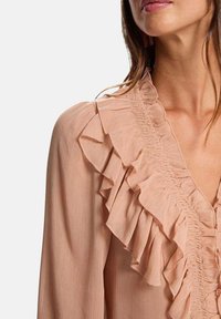 Mujer con blusa rosa claro con detalles de volantes en capas a lo largo del escote en V y los hombros, sobre un fondo liso.