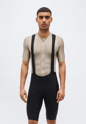 Mann trägt beige Kurzarm-Radtrikot mit Reißverschluss und schwarze Trägerhose vor weißem Hintergrund.