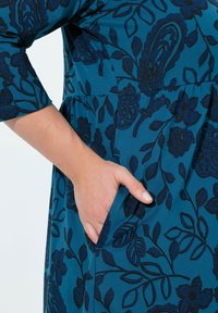 Robe teal avec un motif floral foncé, manches 3/4 et poches latérales. Tissu doux ; coutures visibles au niveau de la taille et de l'ourlet des manches.