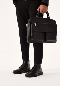 Maletín de cuero negro con dos asas, bolsillo frontal con cremallera, textura suave y un diseño minimalista. Combinado con pantalones y botas negras.