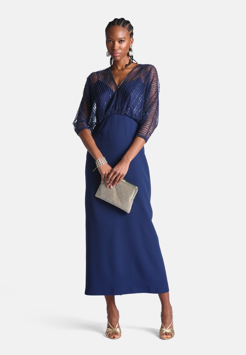 Femme portant une robe maxi bleu marine avec des manches transparentes ornées de perles, tenant une pochette dorée et portant des sandales à talons hauts dorées.