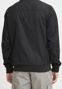 Veste bomber noire avec une texture lisse et brillante, poignets et ourlet côtelés, design simple ; vue de dos montrant une silhouette épurée.