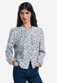 Bluse mit langen Ärmeln, weißem Hintergrund, blauem und grünem Blumenmuster, plissiertem Detail am Ausschnitt und durchgehender Knopfleiste.