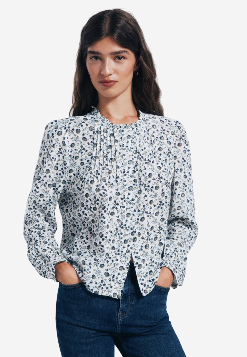 Bluse mit langen Ärmeln, weißem Hintergrund, blauem und grünem Blumenmuster, plissiertem Detail am Ausschnitt und durchgehender Knopfleiste.