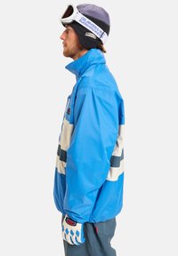 Μπλε αδιάβροχη σακάκι σκι με λευκές και ναυτικές ρίγες, ψηλό κολάρο και μπροστινό φερμουάρ. Ζευγάρι λευκών γαντιών με μπλε διαμαντένιο μοτίβο και γυαλιά.