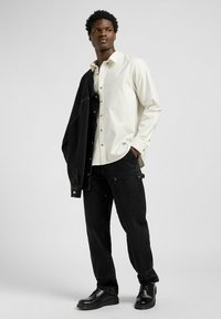 Camisa blanca de botones, chaqueta de mezclilla negra sobre el hombro, jeans ajustados negros y botas negras. Diseño minimalista con botones dorados.
