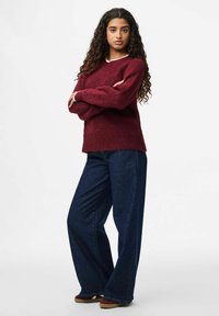 Femme avec de longs cheveux bouclés portant un pull bordeaux, un jean bleu foncé à jambes larges et des chaussures marron, se tenant les bras croisés contre un fond blanc.