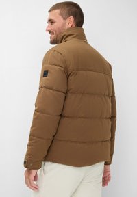 Giacca imbottita marrone con colletto alto, caratterizzata da un design trapuntato e un'etichetta nera del marchio sulla spalla sinistra. Tessuto testurizzato con dettagli a bottoni.