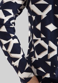 Camisa de manga larga con patrón marino que presenta diseños geométricos en blanco y beige, fabricada con un tejido suave y un corte ajustado.