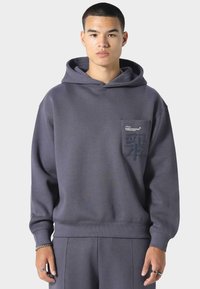 Sudadera gris oscuro hecha de tela suave, con un bolsillo frontal y un diseño de logo texturizado en el bolsillo. Corte holgado con puños de canalé.