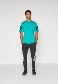 Turkis atletisk T-shirt med sorte tre-striber på ærmerne, kombineret med sorte sweatpants med hvide striber langs siderne.