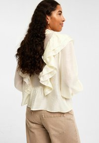 Krämsfärgad blus med volangdetaljer vid axlarna, flödande tyg, långa ärmar och samlad rygg, sliten med beiga byxor.