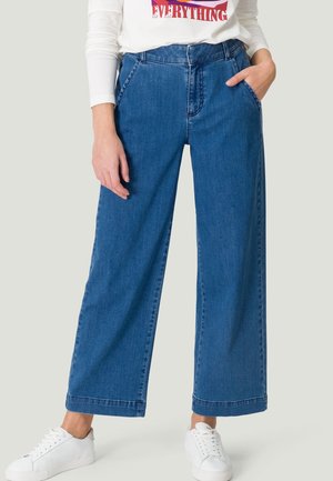 Personne portant un jean bleu à jambes larges, des baskets blanches et un t-shirt blanc à manches longues avec un texte rouge, debout avec une main dans la poche.