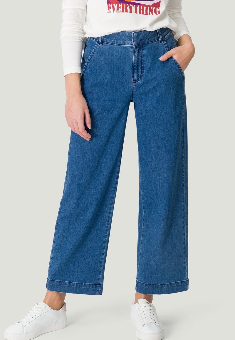 Personne portant un jean bleu à jambes larges, des baskets blanches et un t-shirt blanc à manches longues avec un texte rouge, debout avec une main dans la poche.