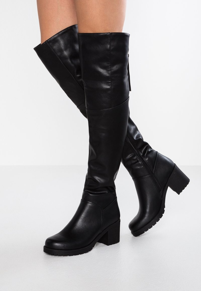 Bottes noires en cuir montantes jusqu'aux genoux avec talons carrés portées sur des jambes croisées sur un fond blanc uni.