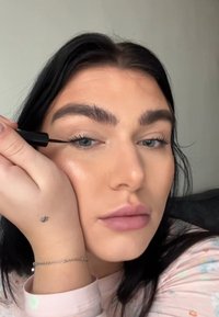 Applicateur de eyeliner liquide noir utilisé sur la paupière ; le sujet a un maquillage léger, de longs cheveux noirs et porte un haut à motif pastel.