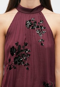 Μπορντό τοπ με halter neck και μαύρες παγιέτες σε σχέδιο λουλουδιών, με απαλή, λεία υφή και ρέον σχέδιο.