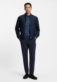 Chaqueta bomber azul marino con cremallera, llevada sobre una camiseta polo azul, combinada con pantalones azul marino oscuros y zapatos de ante negros, posando frente a un fondo blanco.