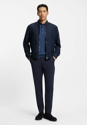 Giubbotto bomber blu marino con zip, indossato sopra una polo blu, abbinato a pantaloni blu scuro e scarpe in camoscio nere, in piedi contro uno sfondo bianco.