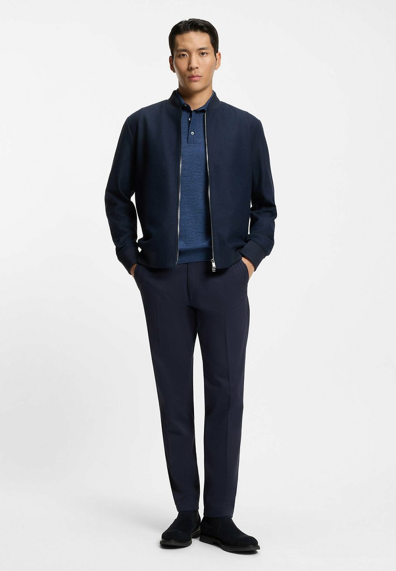 Chaqueta bomber azul marino con cremallera, llevada sobre una camiseta polo azul, combinada con pantalones azul marino oscuros y zapatos de ante negros, posando frente a un fondo blanco.