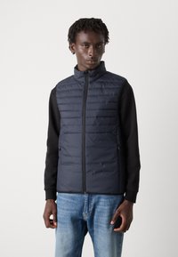 Colete acolchoado azul-marinho com gola alta, fecho de correr e dois bolsos laterais, combinado com uma camisa preta de manga longa e jeans azuis. Textura suave.