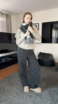 Sweater in light beige, loose black pants, and a black-and-white patterned scarf. Soft-textured beige slippers. Neutral indoor background.