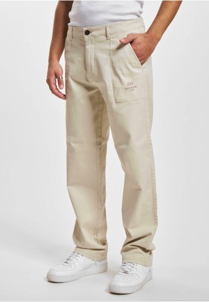 Beige cotton pants with a straight fit, featuring a front pocket and a subtle logo on the side, paired with white sneakers.
