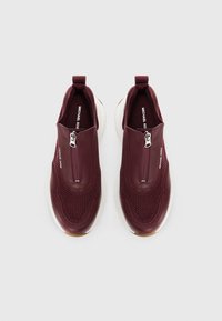 Burgundy slip-on sneakers with breathable mesh, zip closure, and a white rubber sole. Features textured surface and branding inside.