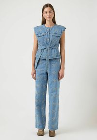 Kobieta ubrana w niebieski denimowy top bez rękawów z paskiem oraz dopasowane haftowane spodnie, stojąca na tle jednolitego jasnego tła.