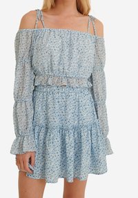 Lyseblå off-shoulder kjole med lange, flæseærmer, indsnævret talje og yderst vidde, der har et lille blomstermønster.