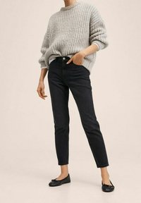 Suéter de punto gris claro con cuello redondo, combinado con jeans ajustados negros. Zapatos planos negros con detalle de lazo. Texturas suaves, corte relajado.