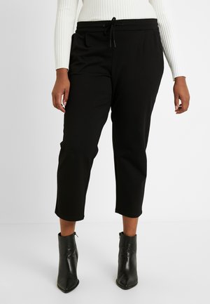 Osoba ubrana w czarne, krótkie spodnie z sznurkiem w pasie i czarne botki na obcasie z ostrym noskiem, stojąca na tle jednolitego tła.