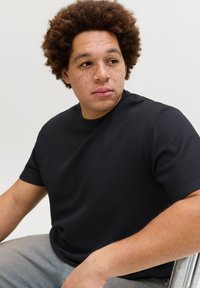 Νέος άνδρας με σγουρά μαλλιά και φακίδες, που φοράει μαύρο κοντομάνικο πουκάμισο, κάθεται και κοιτάζει προς τα πλάγια μπροστά σε απλό φόντο.