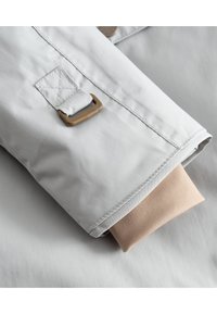 Manica di giacca grigio chiaro con risvolto beige, dotata di accessori color testa di moro e dettagli cuciti, su una superficie liscia.