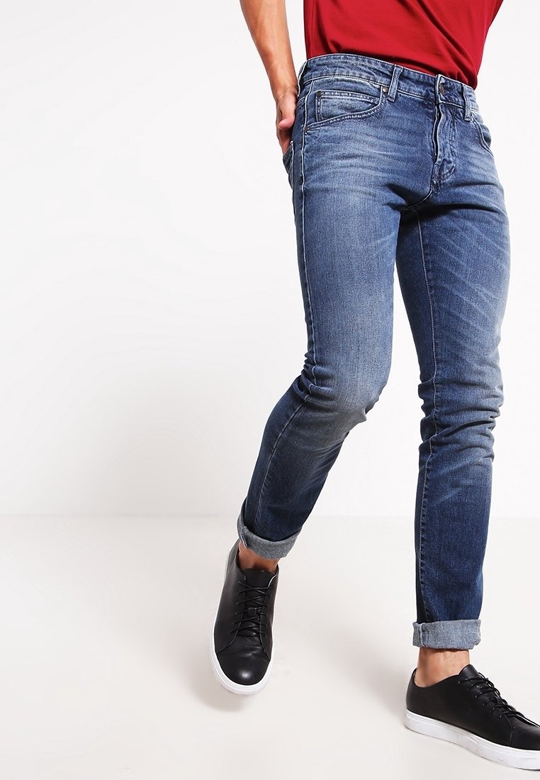 Jean en denim bleu coupe slim avec un lavage délavé, design à cinq poches et revers roulés, porté avec des baskets noires.