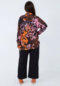 Woman with long hair wears a vibrant, swirling patterned blouse in orange, pink, and black, paired with wide black pants and orange heels.