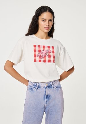 Vrouw met lang donker haar staat met handen in de zij, draagt een wit T-shirt met een roze kreeft op een rood geruit patroon en lichtblauwe spijkerbroek.