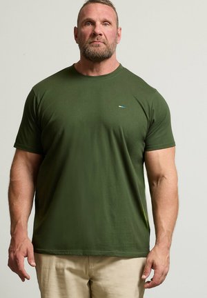 Muskelmand med kort hår og skæg iført en olivengrøn, kortærmet t-shirt og beige bukser, stående mod en ensfarvet baggrund.