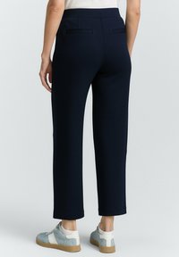Pantaloni blu navy dal taglio sartoriale, con una superficie liscia, tasche posteriori e indossati con sneakers grigie. Dettagli di design minimalisti.