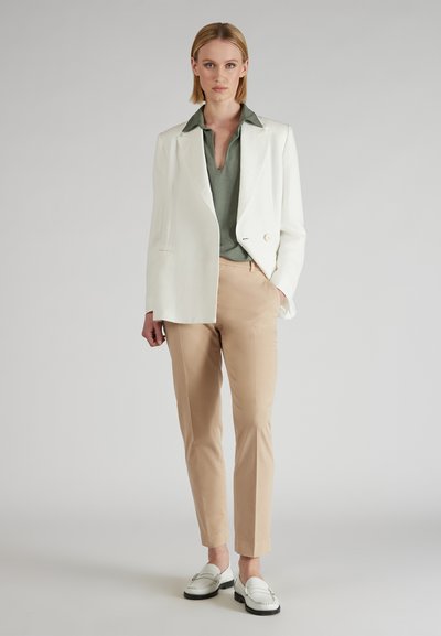 Kobieta stoi w białym żakiecie, zielonej koszuli z kołnierzykiem, beżowych spodniach i białych mokasynach na tle szarego tła.