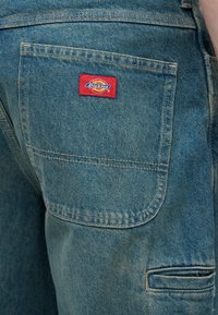 Ανοιχτό μπλε τζιν παντελόνι με μεγάλη τσέπη-patch και κόκκινη ετικέτα Dickies στο πίσω μέρος. Το ύφασμα έχει ελαφρά ξεθώριασμα και κλασική κοπή.