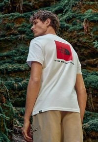 Camiseta de algodón blanca con un logo rojo cuadrado en la parte trasera y texto negro. Mangas cortas, corte holgado, combinada con pantalones cortos beiges.
