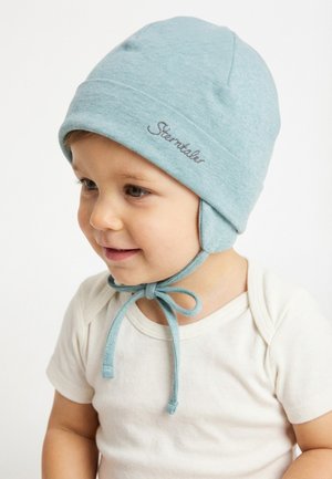 Kleinkind trägt eine hellblaue Mütze mit Ohrklappen, die unter dem Kinn gebunden ist, und ein weißes kurzärmliges Hemd, blickt leicht nach links und lächelt.
