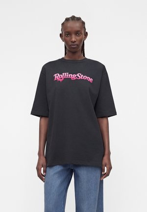 Νεαρό άτομο με κοτσίδες, φορώντας μια μαύρη φαρδιά μπλούζα με ροζ γράμματα "Rolling Stone" και φαρδιά μπλε τζιν, σε απλό φόντο.