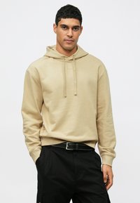 Uomo che indossa una felpa beige con cappuccio e lacci, pantaloni neri, in piedi con una mano in tasca contro uno sfondo bianco.