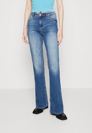 Femme portant un jean flare bleu taille mi-haute avec des cuisses décolorées, un haut sans manches bleu clair et des chaussures noires à bout carré sur un fond blanc uni.