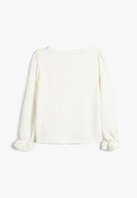 Top blanco de manga larga con tejido texturizado y un patrón de puntos sutil, y puños con fruncidos. Cuello redondo sencillo, sin herrajes visibles.