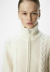 Weißer Strickpullover mit hohem Kragen, vorderem Reißverschluss, strukturiertem Zopfmuster an den Seiten und einer weichen, flauschigen Oberfläche.