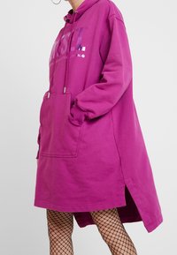 Person som bär en lysande lila oversized huvtröja med framficka och silvertext, tillsammans med svarta fiskenätsstrumpor mot en neutral bakgrund.